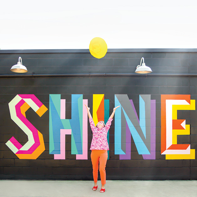 A woman stands in front of a mural that reads "SHINE" in a shot from See Them Shine