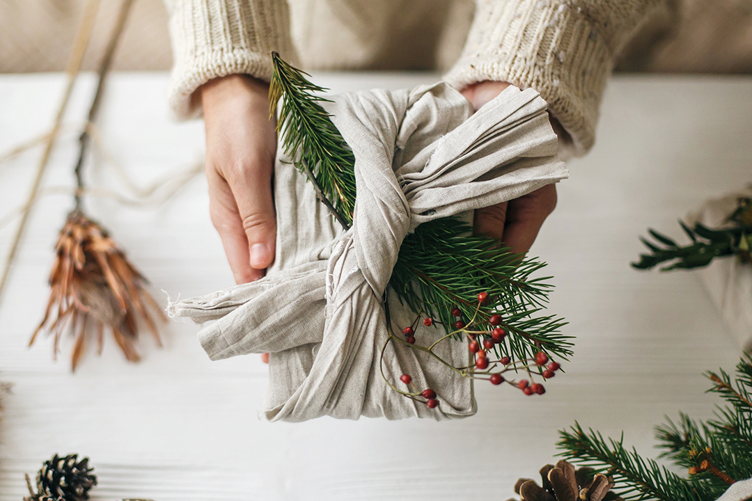 Zero waste Christmas holidays. Hands holding stylish gift wrapped in linen fabric with green fir branch and red berries on rustic background. Plastic free sustainable lifestyle