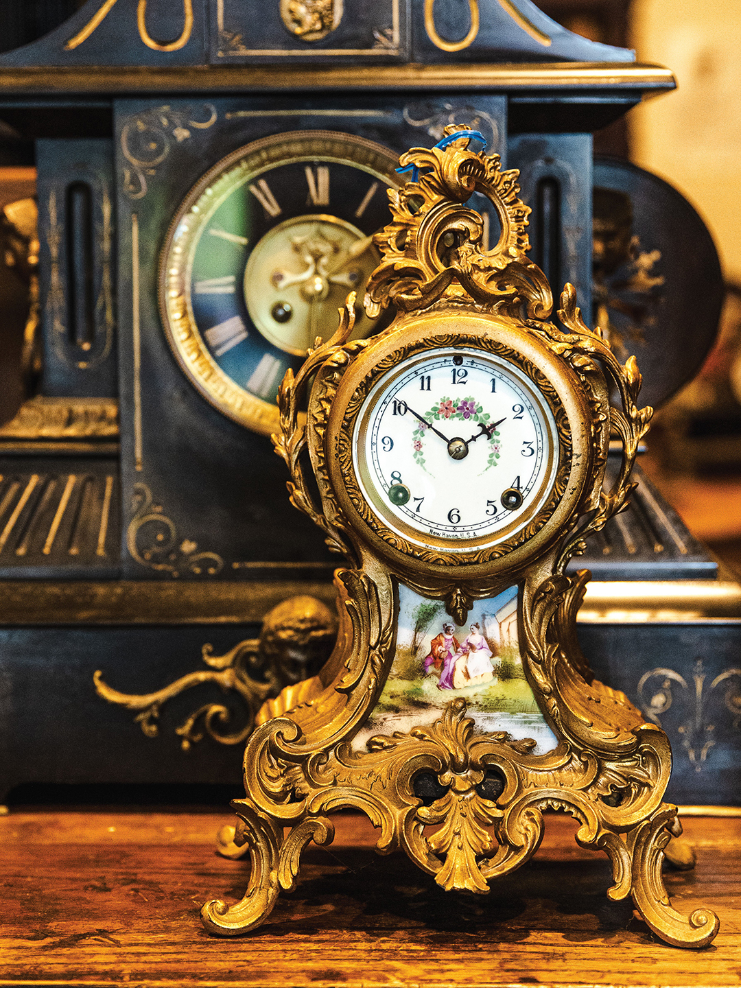 A French mantel clock stands elegantly in front of an antique table clock at Blackstone Manor Clock Repair.